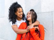 Felice fidanzate multietniche in abiti alla moda in piedi e abbracciandosi contro il muro grigio sulla strada della città di giorno — Foto stock