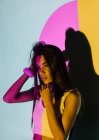 Mujer hispana joven de moda con el brazo levantado mirando hacia otro lado contra la sombra y las luces coloridas del proyector sobre fondo gris - foto de stock