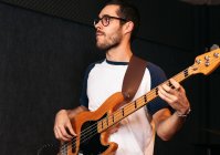 Серйозний молодий хлопець в повсякденному вбранні і окулярах грає на бас-гітарі в легкому клубі — стокове фото