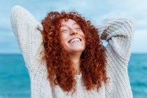Delighted female with curly long ginger hair wearing knitted sweater standing with hands behind head and closed eyes against blue sea — Stock Photo