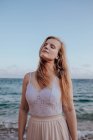 Jovem fêmea atraente com cabelos longos e olhos fechados enquanto estava em pé na praia no verão no fundo borrado — Fotografia de Stock