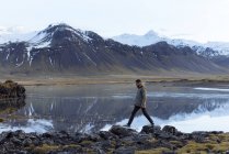 Side view of young male traveler in warm clothes walking along stony coast of calm lake surrounded by snowy mountains during trip in Iceland — Stock Photo