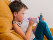 Side view of content sick boy using respirator and breathing oxygen from inhaler while sitting on sofa at home — Stock Photo