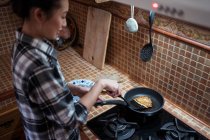 Desde arriba vista lateral de la joven asiática enfocada en ropa casual cocinando deliciosos panqueques usando espátula mientras está de pie en la cocina - foto de stock