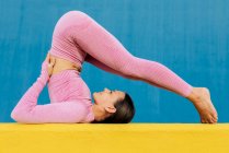 Vista lateral de la hembra delgada flexible en ropa deportiva haciendo ejercicio sobre alfombra amarilla contra la pared azul brillante - foto de stock