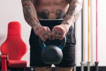 Мощный бодибилдер без рубашек с татуировками, выполняющий упражнения с тяжелыми гирями во время функциональных тренировок в тренажерном зале — стоковое фото
