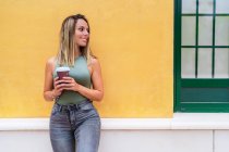 Позитивна жінка з паперовою чашкою кави, посміхаючись і дивлячись на камеру, стоячи біля будівлі на вулиці міста — стокове фото