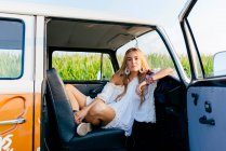 Linda chica rubia sentada dentro de una furgoneta vintage y apoyada en el salpicadero en un día soleado - foto de stock