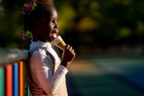 Vista laterale di allegra ragazza afroamericana in piedi vicino a recinzione colorata e leccare dolce gelato sulla strada contro sfondo sfocato — Foto stock