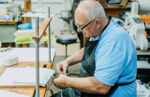 Side view of attentive senior male artisan in apron and eyeglasses tying tapes on wooden board before working on printing press machine — Stock Photo
