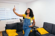 Happy African American female blogger with long curly hair in yellow t shirt and jeans overall standing with hand in pocket and taking selfie on smartphone in light room — Stock Photo