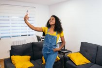 Heureuse blogueuse afro-américaine aux longs cheveux bouclés en t-shirt jaune et jean debout avec la main dans la poche et prenant selfie sur smartphone dans la pièce lumineuse — Photo de stock