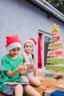 Kinder in roten Nikolausmützen surfen mit dem Handy, während sie in der Nähe von dekorativ bemalten Weihnachtsbaum im hellen Raum während der Weihnachtsfeier sitzen — Stockfoto