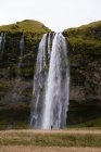 Обратный вид на анонимного путешественника в теплой одежде стоящего и любуясь живописным быстрым водопадом Seljalandsfoss под облачным небом в Исландии — стоковое фото