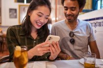 Young Asian female showing photos on smartphone to ethnic boyfriend sitting at table in restaurant — Stock Photo