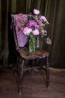 Bouquet de pivoines colorées fraîches et de chrysanthèmes dans un vase en verre placé sur une chaise en bois altérée près de rideaux dans une pièce lumineuse — Photo de stock