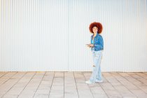 Full body of trendy female with Afro hairstyle wearing stylish outfit with sunglasses browsing cellphone while standing on street near wall — Stock Photo