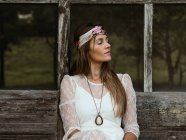 Mujer hippie con los ojos cerrados sentada en el porche de su choza mirando hacia otro lado - foto de stock