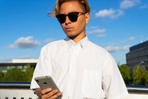 Confiant jeune homme en chemise blanche messagerie texte sur téléphone portable tout en se tenant dans la rue contre le ciel bleu le jour ensoleillé — Photo de stock