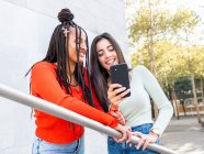 From below of cheerful diverse females in stylish clothes standing on stairs near railing and laughing while browsing smartphone on street in daytime — Stock Photo