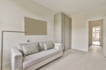 Canapé confortable avec coussins et placards de style minimaliste situé près des fenêtres dans le salon ensoleillé — Photo de stock