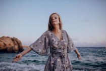 Зачарована жінка з довгим волоссям в модному платті, що стоїть з закритими очима на березі моря в літній вечір — стокове фото