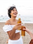 Lieto etnico femminile clinking bottiglia di birra con amico raccolto mentre godendo serata estiva sulla spiaggia di sabbia — Foto stock
