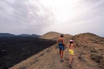 Rückenansicht einer anonymen Mutter mit einem Mädchen, das Händchen hält, während sie in malerischem Blick auf den Krater des Vulkans Caldereta gegen Berge und Meer unter hellem Himmel auf Lanzarote spaziert — Stockfoto