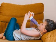 Vista lateral do menino com asma usando o inalador enquanto estava deitado no sofá em casa — Fotografia de Stock