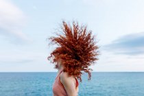 Vista lateral de despreocupada hembra anónima sacudiendo el pelo rizado de jengibre en la costa del mar azul - foto de stock