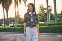 Серьёзная азиатка в современных белых наушниках смотрит вдаль, стоя рядом с дорогой на улице города с зелеными деревьями — стоковое фото