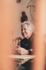 Donna anziana sorridente che indossa vestiti caldi seduta a tavola con tablet e tazza di tè distogliendo lo sguardo — Foto stock