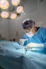 Cirujano profesional senior en máscara y uniforme haciendo operación bajo lámpara en quirófano - foto de stock
