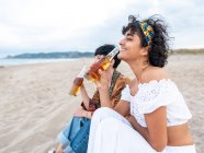 Positivo multietnica migliori amiche bere birra fredda mentre seduto sulla spiaggia di sabbia e ridere in estate sera — Foto stock