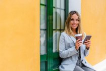 Mujer positiva en traje elegante con café para llevar mirando a la cámara mientras mensajería de texto en el teléfono celular cerca del edificio con bolso - foto de stock