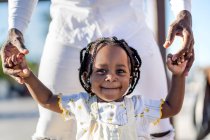 Feliz niña afroamericana en vestido ligero cogida de la mano de un padre irreconocible mientras camina por la calle en un día soleado - foto de stock