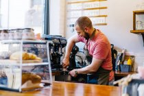 Vue latérale du barista masculin dans le tablier avec portafilter brassant du café à l'aide d'une machine à café tout en se tenant près du comptoir dans le café — Photo de stock
