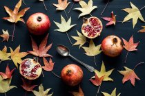 Top view composition of dried autumn leaves placed near plates of ripe sweet pomegranates on black background — Stock Photo
