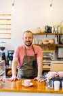 Cheerful male barista in apron smiling and looking at camera while standing at counter with cup of coffee in coffeehouse — Stock Photo