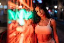 Mulher encantadora em top branco olhando para a câmera enquanto está perto do edifício com luzes brilhantes à noite na rua — Fotografia de Stock