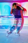 Vista lateral da moda jovem Dominicana millennial feminino com longas tranças afro de pé no chão e olhando para baixo enquanto ouve música em fones de ouvido em sala com iluminação geométrica colorida — Fotografia de Stock