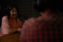 Sonríe joven presentadora de radio femenina con auriculares sentados en la mesa con micrófono y comunicándose con un colega anónimo durante la grabación del podcast en el estudio - foto de stock