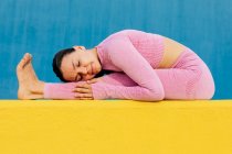 Молодая женщина в розовой спортивной одежде сидит с закрытыми глазами и растягивает ноги на желтом коврике — стоковое фото
