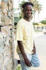 Positivo afro-americano jovem do sexo masculino com o sorriso dos dentes vestindo roupas casuais em pé e inclinado na perna do joelho na parede de pedra — Fotografia de Stock