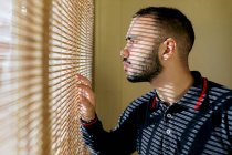 Crop side view of young bearded ethnic male wearing fireman sweat jacket looking out window and touching blinds with fingers in bright sunlight — Stock Photo