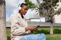 Tranquil African American female with closed eyes listening to music in wireless headphones while sitting on lawn near tree trunk in sunny park while using the smartphone — Stock Photo