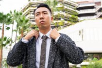 Gut gekleidete junge asiatische männliche Unternehmer mit Krawatte schauen weg, während sie auf der Straße gegen moderne Gebäude in der Stadt bummeln — Stockfoto