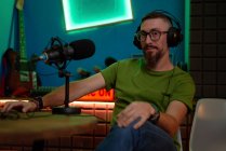 Positive young bearded male millennial in eyeglasses and headphones and talking in mic while recording podcast in dark studio — Stock Photo