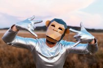 Pessoa irreconhecível com máscara de macaco geométrico demonstrando gesto de rocha em fundo turvo de terra rural — Fotografia de Stock