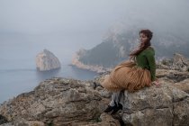 Full body side view of dreamy female in old fashioned clothes sitting on edge of stony cliff near sea in misty weather — Stock Photo